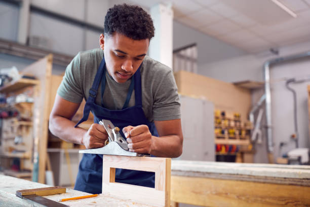 Male Student Studying For Carpentry Apprenticeship At College Using Wood Plane